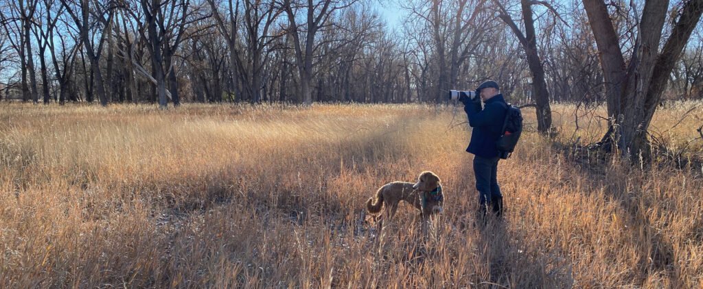 A man and his dog standing in a field of dry grass taking a picture with a long camera lens.